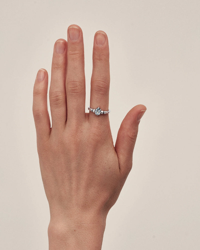 A silver ring with a small cluster of clear gemstones worn on the ring finger of a hand, set against a plain light beige background.