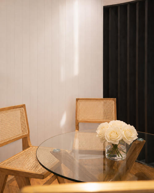 Dining area with a glass table and wicker chairs, featuring a vase of white flowers.